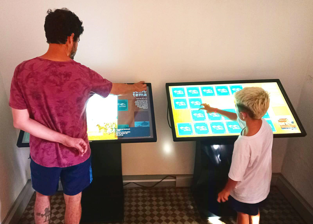 Pai e filho estão em uma sala do museu jogando jogo da memória em telas touch de tv.